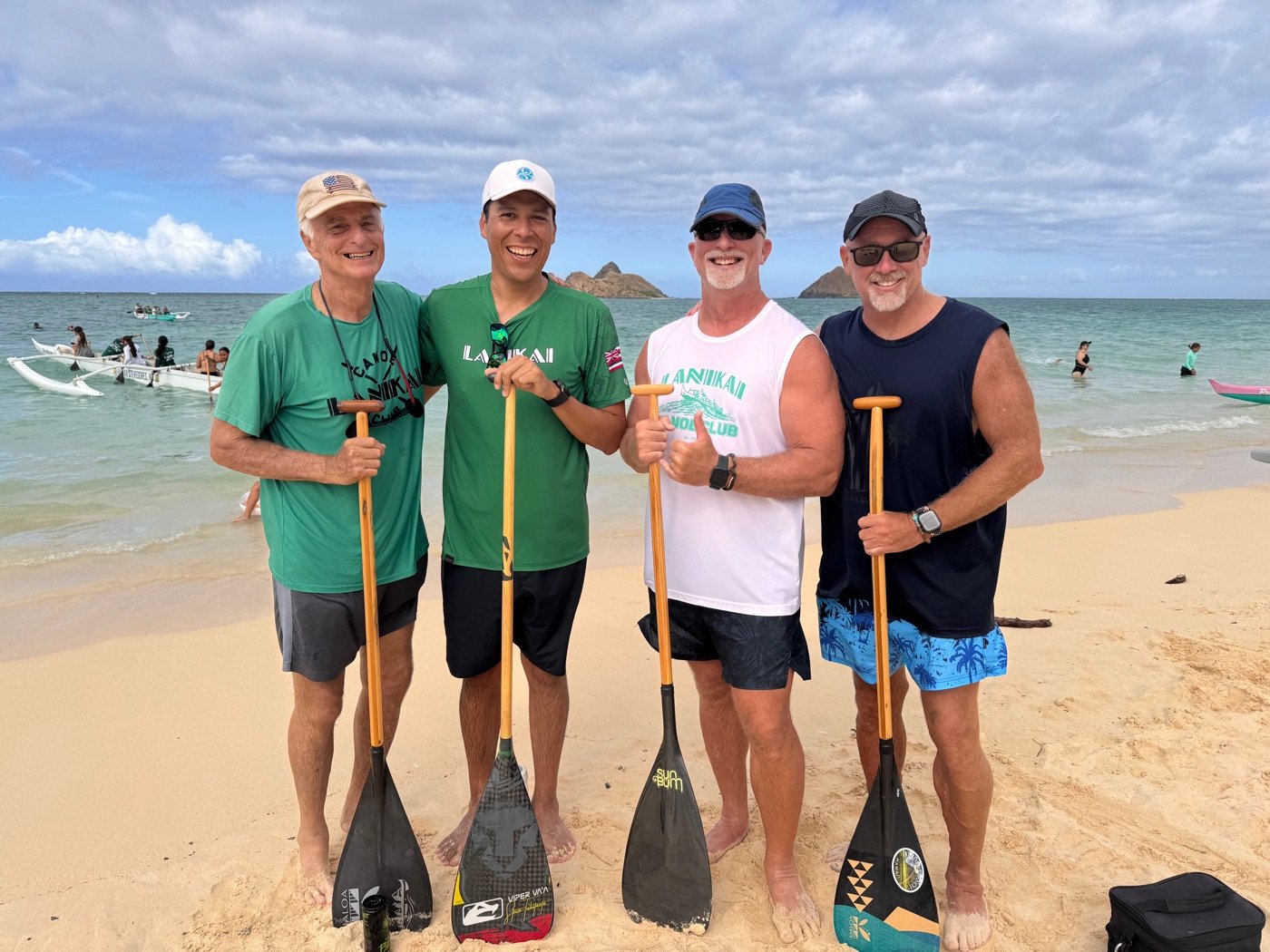 Paul Petro and three fellow Lanikai Canoe Club paddlers on the beach at Lanikai, Oahu, holding their paddles with the Mokulua islands behind them
